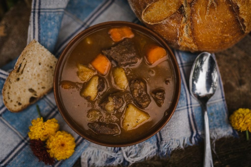 Bowl of brothy beef and vegetable stew with bread and a spoon on a blue cloth