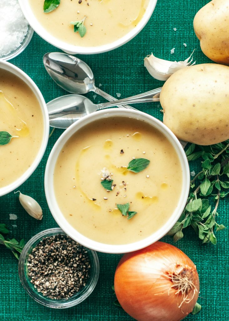 Bowls of smooth potato soup on a green cloth with whole potatoes, onion and black pepper