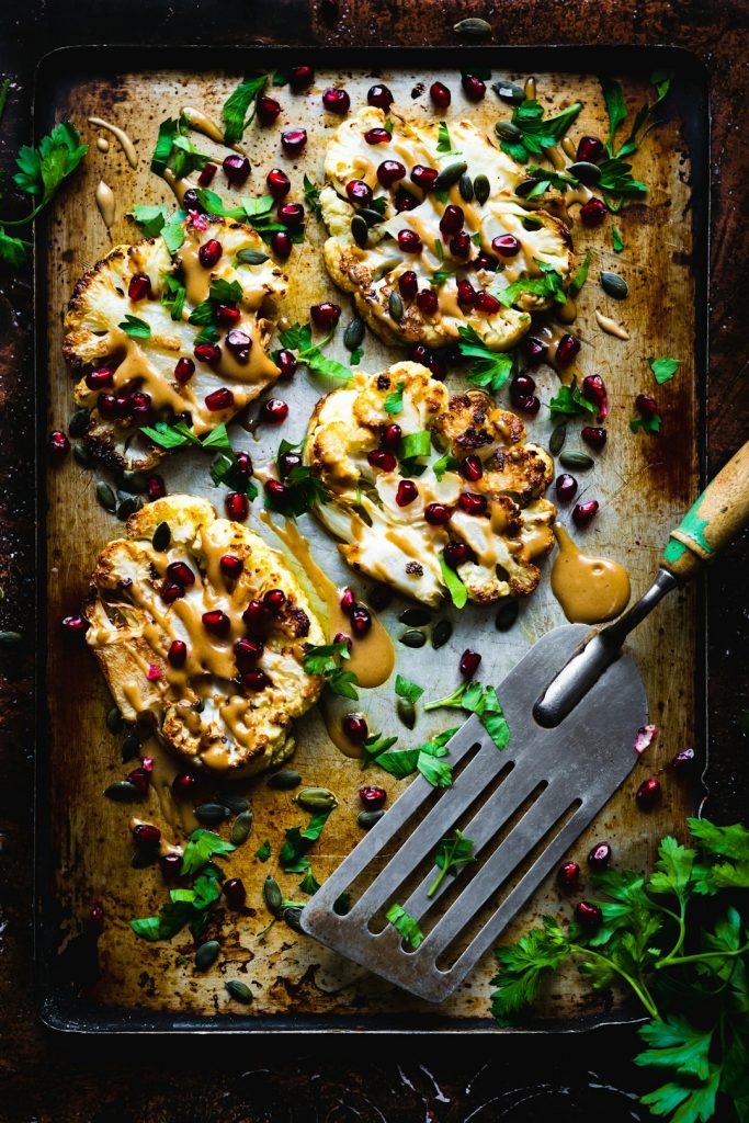 Roasted cauliflower steaks on a sheet pan drizzled with sauce and topped with pomegranate seeds and herbs