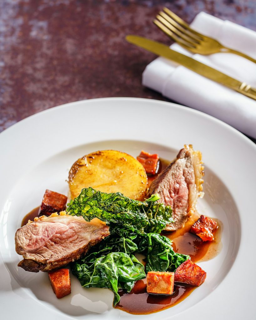 Sliced roast duck breast with a dark fruit sauce, greens and roasted vegetables on a white plate